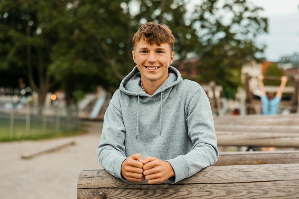 Porträtt av en leende tonårspojke i grå hoodie i en park